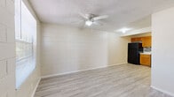 This single-floor unit is built with painted concrete block walls throughout, giving the space a sturdy, consistent character. Light wood-look laminate flooring runs seamlessly across every room, creating a clean and cohesive aesthetic from entry to bedroom.

The open living area is anchored by a ceiling fan with a light fixture and features the distinctive block wall construction painted in a neutral cream tone. A textured popcorn ceiling adds a classic touch overhead. The space flows directly into the kitchen, which is set in a recessed nook and equipped with warm honey-toned wood cabinetry, a mosaic tile backsplash, white countertops, and a full suite of black appliances including a refrigerator and electric range.

The bedroom is generously sized, filled with natural light through a sizable window with blinds, and includes its own ceiling fan. A walk-in closet with multiple shelving tiers provides ample storage space. An additional room off the main living area also features a window and ceiling fan, offering flexible use as a second bedroom, office, or dining space.

The bathroom includes a wood-toned vanity with a marble-style countertop and an integrated sink, alongside a separate water closet with a shower and tub combination. The front entry opens to a bright foyer with a solid front door and an adjacent window, welcoming natural light into the home.