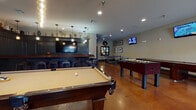 Located at 141 Hemlock Dr in Auburn, Alabama, this single-floor entertainment and game room is designed for social gatherings and recreational enjoyment. The space is anchored by rich, polished concrete floors in warm amber tones that complement the dark wood paneling accented throughout.

At the heart of the room sits a full-service bar with a dark wood facade, a row of cushioned bar stools, pendant lighting, and a large wall-mounted TV — creating an inviting hub for guests. A professional-grade billiards table is prominently positioned beneath an industrial-style brass pendant light fixture, adding a sophisticated focal point to the space.

The game room is further equipped with a foosball table, a shuffleboard table, and a ping pong table, ensuring a wide variety of entertainment options. Multiple wall-mounted flat-screen TVs are strategically placed throughout, making it easy to follow sports from any area of the room.

A lounge nook features dark tufted booth seating, natural light from blinds-covered windows, and decorative shelving displaying vinyl records, framed artwork, and curated collectibles. A striking dragon artwork serves as a bold decorative statement in this area. The overall ambiance blends a classic sports bar aesthetic with a warm, rustic character, making it an ideal venue for group entertainment.