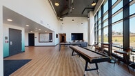 This single-floor space combines contemporary industrial design with a welcoming, social atmosphere. The main lounge area commands attention with its dramatically high ceilings, exposed ductwork, and pendant lighting that lends an urban, loft-inspired character. Warm-toned hardwood-style flooring runs throughout, grounding the open layout and complementing the bright white walls.

A sweeping wall of floor-to-ceiling windows floods the space with abundant natural light and provides expansive outdoor views, creating an airy and energetic environment. The recreational zone is well-appointed, featuring a shuffleboard table, a ping-pong table, and a large wall-mounted flat-screen TV paired with comfortable lounge seating — making it an ideal gathering spot for both casual socializing and organized activities. A framed oversized Scrabble board wall art adds a playful, creative focal point to the lounge area.

Adjacent to the main open area, a stylish semi-private nook is furnished with plush green velvet accent chairs, a round ottoman-style table, and a wall-mounted display screen — offering a more intimate setting for conversation or informal meetings. A glass-walled private room enclosed by sleek black-framed partitions provides a separate, transparent enclosure suitable for small meetings or focused use, maintaining visual connectivity with the broader space while offering a degree of separation.