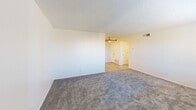 Located at 12400 Montgomery Blvd NE in Albuquerque, New Mexico, this single-story residential unit offers a practical and well-maintained layout suited for comfortable everyday living. The living area is generously sized, featuring plush wall-to-wall carpeting, crisp white walls, and a textured ceiling. A large window fitted with horizontal blinds allows natural light to fill the space while offering views of the surrounding neighborhood. The galley-style kitchen is efficiently designed with white cabinetry lining both sides, grey laminate countertops, a double stainless steel sink, and a patterned tile backsplash. Appliances include an electric range and a stainless steel refrigerator, complemented by wood-look flooring and a flush-mount ceiling light. The bedroom continues the carpeted flooring and neutral color palette, featuring a window with blinds for natural light and a ceiling fan for added comfort. A secondary carpeted room provides additional flexible space. The bathroom is clean and functional, appointed with a white vanity with grey countertop, wood-look tile flooring, a tub-shower combination, and built-in overhead shelving for convenient storage. Throughout the unit, the design emphasizes simplicity, clean finishes, and practical functionality.