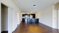 This single-floor residence offers a thoughtfully designed open-concept layout that seamlessly connects the living and kitchen areas, creating a spacious and cohesive atmosphere. Rich, dark hardwood floors extend throughout the space, adding warmth and a sense of continuity.

The kitchen is a standout feature, showcasing dark espresso cabinetry that reaches toward the ceiling, complemented by light-toned countertops that provide a striking contrast. Stainless steel appliances, including a refrigerator and electric range, lend a modern, polished look to the culinary space. A large central island with an integrated sink serves as both a functional workspace and a natural gathering point, with stylish pendant lighting hanging above to illuminate the area.

The open living space adjacent to the kitchen benefits from the generous layout, allowing for flexible furniture arrangements. Crisp white walls and bright ceilings enhance the sense of openness and reflect light evenly across the room. Clean white baseboards provide a finished, tailored look throughout. Doorways leading to additional rooms hint at further private spaces within the home. Overall, the space balances contemporary design elements with a warm, inviting ambiance.