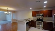 This single-floor space showcases a well-appointed kitchen as its central feature. The kitchen is designed with a functional peninsula that extends from the main counter, providing additional workspace and a natural division between the kitchen and the adjacent living area. The peninsula is topped with speckled granite-style countertops that continue along the main counter run, offering ample prep space.

Rich, dark cherry wood cabinetry lines the upper and lower walls, providing generous storage. The upper cabinets feature sleek bar-style hardware, complementing the modern aesthetic. Stainless steel appliances include an electric range with oven, an over-the-range microwave, and a dishwasher integrated beneath the peninsula, ensuring a cohesive, contemporary look throughout.

The kitchen flows openly into the surrounding living space, which features warm hardwood-style flooring visible in the adjacent area, contrasting with the lighter flooring in the kitchen zone. Recessed lighting in the smooth white ceiling provides even illumination throughout. Neutral-toned walls keep the space feeling bright and open, while white baseboards add a clean finishing detail throughout the room.