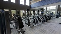 This fitness center offers a spacious, contemporary workout environment designed to support a full range of exercise routines. The room is characterized by soaring ceilings that create an open, airy atmosphere, while a row of large windows along one wall invites abundant natural light and provides views of the surrounding greenery, enhancing the overall workout experience.

The cardio area is prominently featured and well-stocked with a lineup of modern elliptical trainers and treadmills, each equipped with individual display screens for entertainment and performance tracking. A strength training station is also present, adding versatility to the available equipment. The machines are arranged in an organized, spacious layout that allows for comfortable movement throughout the room.

The interior design is sleek and modern, with neutral-toned carpet tiles, gray walls accented by a large decorative graphic panel near the ceiling, and floor-to-ceiling mirrors that visually expand the space and assist with form during workouts. The overall ambiance is clean, professional, and energizing, making it a well-appointed facility suited for a variety of fitness goals.