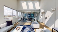 This single-floor amenity space is defined by its striking architectural design, most notably the series of expansive skylights that run across the vaulted ceiling, flooding the interior with abundant natural light throughout the day. Hardwood-style flooring and crisp white walls create a clean, modern aesthetic that ties the various zones together cohesively.

The central lounge area serves as the heart of the space, furnished with contemporary sofas and accent chairs arranged around a bold navy and white geometric rug. A floor-to-ceiling glass wall and wide sliding doors connect the lounge directly to an outdoor pool terrace, offering sweeping city skyline views. A marble-tiled fireplace wall features a built-in flat-screen TV above a linear electric firebox, adding a refined focal point.

Adjacent to the lounge, a generous dining and social area provides multiple wooden tables with navy upholstered chairs, ideal for group gatherings. The fully equipped kitchen features dark wood cabinetry, stainless steel appliances including a professional-grade gas range, and a vibrant teal tile backsplash that adds a bold pop of color.

Enclosed behind glass walls, the dedicated fitness center is well-equipped with cardio machines and benefits from large windows framing city views, creating an energizing workout environment.