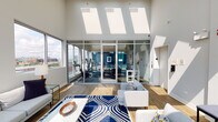 This single-floor amenity space is defined by its striking architectural design, most notably the series of expansive skylights that run across the vaulted ceiling, flooding the interior with abundant natural light throughout the day. Hardwood-style flooring and crisp white walls create a clean, modern aesthetic that ties the various zones together cohesively.

The central lounge area serves as the heart of the space, furnished with contemporary sofas and accent chairs arranged around a bold navy and white geometric rug. A floor-to-ceiling glass wall and wide sliding doors connect the lounge directly to an outdoor pool terrace, offering sweeping city skyline views. A marble-tiled fireplace wall features a built-in flat-screen TV above a linear electric firebox, adding a refined focal point.

Adjacent to the lounge, a generous dining and social area provides multiple wooden tables with navy upholstered chairs, ideal for group gatherings. The fully equipped kitchen features dark wood cabinetry, stainless steel appliances including a professional-grade gas range, and a vibrant teal tile backsplash that adds a bold pop of color.

Enclosed behind glass walls, the dedicated fitness center is well-equipped with cardio machines and benefits from large windows framing city views, creating an energizing workout environment.