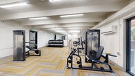 Located at 8680 E Alameda Ave in Denver, Colorado, this fitness center offers a comprehensive workout facility designed to accommodate a variety of exercise routines. The space is laid out across a single floor and features a distinctively patterned herringbone carpet in yellow and gray tones throughout, providing a cohesive and functional aesthetic.

The facility is organized into distinct workout zones. A dedicated cardio area houses multiple treadmills, elliptical trainers, stair climbers, and rowing machines, positioned near large windows that allow natural light and views of the surrounding outdoor greenery. A strength training section features a full dumbbell rack with a wide range of weights, a multi-station cable machine by Precor, a lat pulldown and row machine, a bench press station, and additional selectorized weight machines.

A separate area is set aside for cycling, equipped with several stationary spin bikes, kettlebells, and a rowing machine, accompanied by a mirrored wall to support form and technique. Throughout the facility, large wall-mounted mirrors enhance the sense of space and assist users during workouts. Overhead fluorescent lighting ensures consistent, bright illumination across all zones. The textured ceiling with exposed beam detailing adds a structured architectural character to the space. Multiple entry and exit points with glass-panel doors provide easy access throughout the facility.