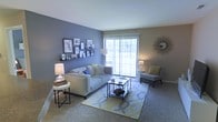 This single-floor residence offers a comfortable and thoughtfully designed living space with a warm, inviting ambiance. The living room serves as the heart of the home, showcasing a combination of cool blue and neutral taupe accent walls that create a calm, modern aesthetic. Plush wall-to-wall carpeting adds a cozy underfoot feel, while a large sliding glass door floods the room with natural light and provides a view to the outdoors, enhancing the sense of openness.

The living area is generously proportioned, offering ample room for both relaxed seating and entertaining. A soft grey sofa anchors the space, complemented by a coordinating accent chair, a glass-and-metal coffee table, and layered side tables that add functional style. A patterned area rug in muted yellow and grey tones grounds the seating arrangement and adds a decorative focal point.

A decorative gallery wall featuring an assortment of framed artwork and photographs is mounted on floating shelves along the blue accent wall, adding a personalized and curated touch. Soft ambient lighting from table lamps on either side of the seating area creates a welcoming glow. A sunburst mirror accent on the adjacent wall adds a finishing decorative detail, contributing to the space's polished and cohesive interior style.