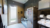 This single-level Pet Spa is a purpose-built grooming facility designed with pet owners in mind. The space centers around a stainless steel dog wash station, providing a hygienic and elevated bathing surface that makes washing pets convenient and efficient. Adjacent to the wash station is a Revolution Dog grooming unit, a professional-grade appliance equipped with animal care instructions and secure lockable storage, ensuring all grooming essentials are organized and readily accessible. Fresh towels are kept on hand nearby, adding a practical touch to the overall setup. The interior walls are clad in dark mosaic tile, lending a sleek, modern aesthetic to the space, while light-toned floor tiles keep the environment bright and easy to clean. Natural light filters in through a glass door that opens to an outdoor area, providing ventilation and a welcoming atmosphere. Informational signage is posted on the walls for guidance during use. The overall design balances functionality with a polished, contemporary look, offering a clean and well-equipped environment for routine pet grooming needs.