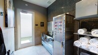 This single-level Pet Spa is a purpose-built grooming facility designed with pet owners in mind. The space centers around a stainless steel dog wash station, providing a hygienic and elevated bathing surface that makes washing pets convenient and efficient. Adjacent to the wash station is a Revolution Dog grooming unit, a professional-grade appliance equipped with animal care instructions and secure lockable storage, ensuring all grooming essentials are organized and readily accessible. Fresh towels are kept on hand nearby, adding a practical touch to the overall setup. The interior walls are clad in dark mosaic tile, lending a sleek, modern aesthetic to the space, while light-toned floor tiles keep the environment bright and easy to clean. Natural light filters in through a glass door that opens to an outdoor area, providing ventilation and a welcoming atmosphere. Informational signage is posted on the walls for guidance during use. The overall design balances functionality with a polished, contemporary look, offering a clean and well-equipped environment for routine pet grooming needs.