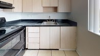 This single-floor unit presents a functional and straightforward layout, as revealed by the 3D floor plan, which shows a living area, kitchen, bedrooms, and bathroom arranged in a compact, easy-to-navigate configuration. The living and dining areas are finished with warm wood-look laminate flooring that extends throughout the main spaces, creating a cohesive and low-maintenance surface. A sliding glass door in the living area opens to an outdoor view of lush greenery, while a pendant light fixture marks the dining space near the front entry, where tile flooring provides a practical transition zone.

The kitchen features light wood cabinetry extending to the upper and lower levels, dark granite-style countertops, a stainless steel sink, and tile flooring. It offers ample storage and a clean, utilitarian design. The bedrooms are carpeted for comfort and are distinguished by their painted concrete block accent walls, lending a distinctive architectural character. Each bedroom includes a window with vertical blinds, allowing natural light and outdoor views.

The bathroom is finished with wood-look flooring and features a full soaking tub with a showerhead, a toilet, and a vanity with dark countertop. A small window above the tub brings in natural light, keeping the space bright and airy.