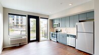 Located at 590 Park St in Saint Paul, Minnesota, this efficiently designed studio unit offers a well-organized single-floor layout comprising an open-plan kitchen and living area, plus one full bathroom. The kitchen is a standout feature, outfitted with sleek blue-gray flat-front cabinetry extending to the ceiling for generous storage, a white subway tile backsplash, light countertops, and a full suite of stainless steel appliances including a refrigerator, electric range, built-in microwave, and dishwasher. Warm-toned hardwood-style flooring runs continuously throughout the entire unit, lending a cohesive and inviting feel. The living area is bright and open, benefiting from large black-framed windows and a glass-panel door that leads directly to a private balcony, offering outdoor views of the surrounding Saint Paul neighborhood. The bathroom features a wide vanity with ample under-sink storage, a large mirror, and matching gray cabinetry consistent with the kitchen's modern aesthetic. A hallway connects the main living space to the bathroom, complete with wall-mounted hooks for added convenience. Recessed lighting and neutral wall tones throughout contribute to a clean, contemporary ambiance that maximizes the sense of space within this thoughtfully designed unit.