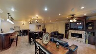 This inviting club room is thoughtfully designed to accommodate a variety of social gatherings and events, offering multiple distinct zones within a single open-plan layout. The lounge area serves as the focal point, anchored by a stone-faced fireplace with a built-in entertainment unit, a wall-mounted flat-screen television, and a ceiling fan overhead. Plush carpeting and rich leather seating — including sofas and accent chairs — create a warm, comfortable atmosphere throughout.

Adjacent to the lounge, a full kitchen bar is outfitted with dark wood cabinetry, stone tile backsplash, stainless steel appliances, and a generous bar counter lined with woven bar stools, making it well-suited for entertaining. Pendant lighting and elegant chandeliers illuminate the space, adding a refined ambiance.

A separate dining area with high-top tables and chairs provides additional seating for guests. Decorative touches such as branch-inspired wall panels and draped curtains contribute to the room's warm, nature-inspired aesthetic. A large projection screen along one wall offers a dedicated space for presentations or movie screenings. French doors allow natural light to filter in, connecting the interior to the outdoors. Crown molding and recessed lighting complete the polished, community-oriented design of this versatile club room.