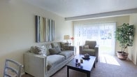 This single-floor space centers around a well-appointed living area designed for comfort and functionality. The living room is finished with neutral-toned walls and a textured ceiling, creating a clean and understated aesthetic. A large sliding glass door with vertical blinds serves as a prominent architectural feature, allowing generous amounts of natural light to fill the room while offering direct access to an outdoor area and views of surrounding greenery.

The sitting area is arranged around a dark wood coffee table and accommodates a plush, light-toned sofa paired with a matching armchair, both positioned to take advantage of the natural light and outdoor view. Decorative accent pillows in earthy tones complement the neutral palette throughout the room. A side table with a classic table lamp and a small floral arrangement adds a warm, residential touch to the space.

Wall décor includes a large contemporary canvas painting in muted greens, blues, and grays, lending a subtle artistic focal point to the room. The carpeted flooring contributes to the overall warmth and comfort of the interior. Together, these elements create a calm, inviting ambiance suited for everyday living.