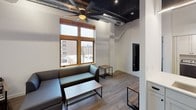 This single-floor unit showcases a compelling blend of industrial and contemporary design elements throughout its well-organized layout. The living area immediately draws attention with its dramatically tall exposed ceiling, complete with visible ductwork and dark-painted beams that lend an authentic loft-like character. A large ceiling fan anchors the space, while wood-framed windows frame urban rooftop views and allow natural light to fill the room. The seating area is furnished with a sectional sofa, a coffee table, and side tables with sleek metal-and-wood construction, reinforcing the industrial aesthetic. Light hardwood-style flooring runs throughout the unit, providing visual continuity.

A bold teal accent column serves as a dividing feature between the living space and kitchen, with a wall-mounted flat-screen television integrated directly into it. The kitchen is outfitted with white shaker cabinetry, white subway tile backsplash, light countertops, and stainless steel appliances including a range, microwave, dishwasher, and refrigerator. In-unit washer and dryer are conveniently accessible nearby.

The bedroom is a compact, functional space with hardwood flooring, a ceiling fan, a bed frame, and a wall-mounted monitor above a wooden desk. The bathroom features clean white finishes, black hardware accents, a vanity with an integrated sink, and recessed lighting.