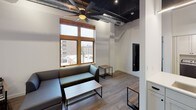 This single-floor unit showcases a compelling blend of industrial and contemporary design elements throughout its well-organized layout. The living area immediately draws attention with its dramatically tall exposed ceiling, complete with visible ductwork and dark-painted beams that lend an authentic loft-like character. A large ceiling fan anchors the space, while wood-framed windows frame urban rooftop views and allow natural light to fill the room. The seating area is furnished with a sectional sofa, a coffee table, and side tables with sleek metal-and-wood construction, reinforcing the industrial aesthetic. Light hardwood-style flooring runs throughout the unit, providing visual continuity.

A bold teal accent column serves as a dividing feature between the living space and kitchen, with a wall-mounted flat-screen television integrated directly into it. The kitchen is outfitted with white shaker cabinetry, white subway tile backsplash, light countertops, and stainless steel appliances including a range, microwave, dishwasher, and refrigerator. In-unit washer and dryer are conveniently accessible nearby.

The bedroom is a compact, functional space with hardwood flooring, a ceiling fan, a bed frame, and a wall-mounted monitor above a wooden desk. The bathroom features clean white finishes, black hardware accents, a vanity with an integrated sink, and recessed lighting.