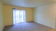 This single-floor unit features a open living area finished with wall-to-wall carpeting in a neutral tone, creating a comfortable and cohesive foundation throughout the space. The room is bathed in natural light, courtesy of a full-width sliding glass door fitted with sheer vertical blinds that lead out to a private balcony. The balcony provides views of the surrounding neighborhood, adding an outdoor extension to the living area. The walls are painted in a soft, warm yellow, lending a bright and welcoming atmosphere to the interior. The smooth white ceiling is clean and unadorned, contributing to the overall sense of openness. Electrical outlets are conveniently positioned along the walls, offering practical functionality. The sliding door arrangement not only maximizes light penetration but also creates a strong visual connection between the interior and the outdoor space, making the room feel more expansive than its footprint suggests. Overall, the space presents a clean, move-in-ready environment with a simple, functional layout suited to a variety of furnishing arrangements.