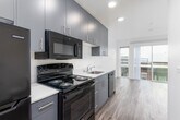This single-floor unit showcases a clean, contemporary design characterized by crisp white walls, recessed lighting, and wood-look flooring that runs consistently throughout the space. A central hallway connects the various areas of the unit, with sliding barn-style doors adding a modern touch while maximizing space efficiency.

The kitchen is thoughtfully designed with a galley-style layout, featuring floor-to-ceiling dark gray cabinetry that provides ample storage. White countertops offer a striking contrast against the cabinetry, while black appliances — including a gas range, microwave, dishwasher, and refrigerator — complete the functional setup. A stainless steel sink anchors the countertop workspace.

Conveniently tucked near the kitchen is a dedicated in-unit laundry area housing a stacked washer and dryer unit, adding a practical and sought-after amenity.

The living area is a standout feature, bathed in natural light through large floor-to-ceiling sliding glass doors that open to views of the surrounding neighborhood streetscape. The open feel of this room is enhanced by its clean lines and neutral palette.

The bathroom maintains the unit's modern aesthetic with simple white fixtures, chrome hardware, and wood-look flooring, keeping the overall design cohesive throughout the space.