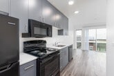 This single-floor unit showcases a clean, contemporary design characterized by crisp white walls, recessed lighting, and wood-look flooring that runs consistently throughout the space. A central hallway connects the various areas of the unit, with sliding barn-style doors adding a modern touch while maximizing space efficiency.

The kitchen is thoughtfully designed with a galley-style layout, featuring floor-to-ceiling dark gray cabinetry that provides ample storage. White countertops offer a striking contrast against the cabinetry, while black appliances — including a gas range, microwave, dishwasher, and refrigerator — complete the functional setup. A stainless steel sink anchors the countertop workspace.

Conveniently tucked near the kitchen is a dedicated in-unit laundry area housing a stacked washer and dryer unit, adding a practical and sought-after amenity.

The living area is a standout feature, bathed in natural light through large floor-to-ceiling sliding glass doors that open to views of the surrounding neighborhood streetscape. The open feel of this room is enhanced by its clean lines and neutral palette.

The bathroom maintains the unit's modern aesthetic with simple white fixtures, chrome hardware, and wood-look flooring, keeping the overall design cohesive throughout the space.