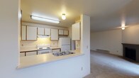 This single-floor residence offers a practical and well-organized layout across multiple connected living areas. The kitchen is designed with functionality in mind, featuring white cabinetry with wood-trimmed accents, a white electric range, a full-size refrigerator, and an under-cabinet range hood. A peninsula counter with a double-basin stainless steel sink extends into the space, creating a natural division between the kitchen and the adjoining living area while offering additional prep and serving space. The living area is generously sized, finished with neutral wall-to-wall carpeting, and anchored by a tile-surround fireplace that serves as a focal point. Baseboard heating runs along the walls throughout the unit, providing consistent climate control. A ceiling fan adds to the comfort of the main living space. Two carpeted bedrooms each feature large windows with horizontal blinds that allow natural light and views of surrounding greenery. Ample closet storage is available, including a double-door closet visible from the main living space. The bathroom includes a white vanity with a single-basin sink, mirrored medicine cabinets for storage, tile flooring, and a full-size soaking tub with a shower. The overall color palette is light and neutral, creating a clean, open atmosphere throughout.