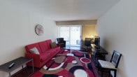 This single-floor space combines comfort and functionality in a bright, inviting layout. The main living area is the centerpiece of the space, immediately drawing attention with its bold, colorful area rug featuring circular patterns in red, black, and beige tones. A striking red sofa anchors the seating area alongside a dark accent chair, creating a lively and welcoming atmosphere. A decorative wall clock adds a casual, modern touch to the neutral-toned walls.

Natural light fills the room through a large sliding glass door fitted with vertical blinds, which opens to a private outdoor balcony — a standout feature that enhances the sense of openness and connection to the outdoors. The dedicated workspace along the far wall includes a sleek dark desk paired with an ergonomic office chair and a monitor setup, making the space well-suited for both relaxing and remote work.

The flooring beneath the rug appears to be a dark hardwood or laminate, adding warmth and contrast to the lighter walls and ceiling. Overall, the space presents a compact yet efficiently utilized layout with a vibrant, contemporary character that balances leisure and productivity seamlessly.