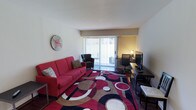 This single-floor space combines comfort and functionality in a bright, inviting layout. The main living area is the centerpiece of the space, immediately drawing attention with its bold, colorful area rug featuring circular patterns in red, black, and beige tones. A striking red sofa anchors the seating area alongside a dark accent chair, creating a lively and welcoming atmosphere. A decorative wall clock adds a casual, modern touch to the neutral-toned walls.

Natural light fills the room through a large sliding glass door fitted with vertical blinds, which opens to a private outdoor balcony — a standout feature that enhances the sense of openness and connection to the outdoors. The dedicated workspace along the far wall includes a sleek dark desk paired with an ergonomic office chair and a monitor setup, making the space well-suited for both relaxing and remote work.

The flooring beneath the rug appears to be a dark hardwood or laminate, adding warmth and contrast to the lighter walls and ceiling. Overall, the space presents a compact yet efficiently utilized layout with a vibrant, contemporary character that balances leisure and productivity seamlessly.