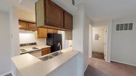 Located at 1956 Chimney Ln. in Kettering, Ohio, this single-floor residence offers a practical and comfortable layout designed for everyday living. The kitchen is well-equipped with warm wood-toned cabinetry, a black refrigerator, an electric range, and a double stainless steel sink set into a peninsula-style breakfast bar that opens toward the living area. Wood-look plank flooring runs through the kitchen and entry hallway, adding a clean, durable finish to the functional space. The living room is generously sized, featuring soft carpeting and a large sliding glass door with vertical blinds that floods the room with natural light and provides access to an outdoor area. The open sightline from the living room to the kitchen creates a connected, airy feel throughout the main living space. The bedroom is a well-proportioned, carpeted room with a window that brings in natural light and includes an adjacent door leading to additional storage or a closet. Neutral wall tones are consistent throughout, lending a calm and versatile backdrop. A hallway connects the main living areas to the bathroom, which features a vanity with matching wood cabinetry and built-in lighting. The overall layout is efficient, with a clear circulation path and practical finishes suited for comfortable daily living.