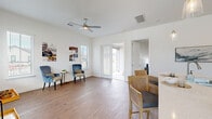 This single-story home offers a clean, modern aesthetic throughout, characterized by crisp white walls, wide-plank wood-look flooring, and an abundance of natural light streaming through large windows fitted with horizontal blinds. The open living area is generously proportioned, anchored by a ceiling fan and connected directly to the outdoors via glass patio doors, creating a seamless indoor-outdoor flow. A dedicated sitting nook, bathed in corner window light, provides a quiet retreat ideal for relaxation or a home office setup. The kitchen is a functional centerpiece, featuring warm wood cabinetry, a classic tile backsplash, stainless steel appliances including a refrigerator with ice maker and a microwave, and a large island with quartz countertops and bar seating — perfect for casual dining or entertaining. Adjacent to the kitchen, the dining area is bright and inviting, with natural light casting soft patterns across the wood-look floors. The bedroom is carpeted for comfort and includes window access and proximity to additional rooms. The covered front porch extends the living space outdoors, offering a relaxed seating area overlooking a manicured lawn and a white picket-fenced yard with pleasant neighborhood views.