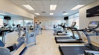 This fitness center is a spacious, well-appointed facility designed to support a comprehensive workout experience. The space is finished with light tile flooring throughout and features a drop panel ceiling with recessed fluorescent lighting, creating a bright and clean environment. Large windows along one wall allow natural light to pour in while offering views of the surrounding urban cityscape, adding an energizing backdrop to workouts.

The cardio section is well-stocked with True-brand treadmills, upright and recumbent stationary bikes, and elliptical machines, all positioned to maximize space and usability. Multiple flat-screen televisions are mounted throughout the room, providing entertainment options during exercise. The strength training area features a robust selection of Cybex and other cable and resistance machines arranged across the floor, accommodating a wide range of muscle groups and exercise styles.

Full-length mirrors line a significant portion of the walls, visually expanding the space and allowing users to monitor their form. A dedicated storage rack with rolled towels is conveniently accessible, and a water station is available nearby. The overall ambiance is clean, functional, and professional, reflecting a well-maintained facility suited for both light and intensive fitness routines.