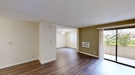 This single-floor residence offers a well-organized layout with a seamless flow between its primary living spaces. The main living area features rich, dark hardwood-style flooring, neutral cream-toned walls accented by an olive green feature wall, and a textured ceiling. A wall-mounted air conditioning unit provides climate control, while a sliding glass door fills the space with natural light and provides access to an outdoor area with lush greenery visible beyond. The open layout connects naturally to an additional room, illuminated by a ceiling fan with a light fixture and a bright window with vertical blinds.

The kitchen is efficiently designed with white cabinetry trimmed in warm wood accents, granite-style countertops, and a full suite of white appliances including a range, microwave, dishwasher, and refrigerator. A recessed ceiling detail and built-in lighting add a subtle architectural touch to the space.

An adjoining dining area features the same hardwood-style flooring, a ceiling fan overhead, and direct sightlines to the kitchen, making it ideal for everyday living and entertaining.

The bathroom is neatly finished with hardwood-style flooring, a wood-trimmed vanity with a granite-style countertop, a large mirror, and bright overhead vanity lighting, creating a clean and functional space throughout.