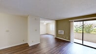 This single-floor residence offers a well-organized layout with a seamless flow between its primary living spaces. The main living area features rich, dark hardwood-style flooring, neutral cream-toned walls accented by an olive green feature wall, and a textured ceiling. A wall-mounted air conditioning unit provides climate control, while a sliding glass door fills the space with natural light and provides access to an outdoor area with lush greenery visible beyond. The open layout connects naturally to an additional room, illuminated by a ceiling fan with a light fixture and a bright window with vertical blinds.

The kitchen is efficiently designed with white cabinetry trimmed in warm wood accents, granite-style countertops, and a full suite of white appliances including a range, microwave, dishwasher, and refrigerator. A recessed ceiling detail and built-in lighting add a subtle architectural touch to the space.

An adjoining dining area features the same hardwood-style flooring, a ceiling fan overhead, and direct sightlines to the kitchen, making it ideal for everyday living and entertaining.

The bathroom is neatly finished with hardwood-style flooring, a wood-trimmed vanity with a granite-style countertop, a large mirror, and bright overhead vanity lighting, creating a clean and functional space throughout.