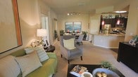 This single-floor residence offers a thoughtfully designed open-concept layout that seamlessly connects the living, dining, and kitchen areas. The living room is a welcoming space anchored by plush seating, including a green sofa and a classic wingback armchair with nailhead trim, all set atop warm neutral carpeting. A large decorative mirror enhances the sense of space, while a table lamp adds a soft, ambient glow. Track lighting overhead provides flexible illumination across the open floor plan. The dining area flows naturally from the living space, featuring a round table surrounded by upholstered chairs and accented by vibrant artwork depicting serene landscape scenes. Lush potted plants are positioned throughout, bringing touches of greenery and life to the interior. Just beyond, the kitchen is partially visible through a raised breakfast bar with granite-style countertops, complemented by pendant lighting that adds a warm, inviting ambiance. The overall color palette consists of soft beiges and creams, creating a cohesive and calming atmosphere throughout. The layout is functional yet elegant, making excellent use of the available space while maintaining an open and airy feel.