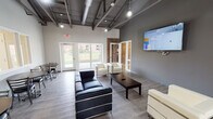 Located at 1958 W Algonquin Road in Mount Prospect, Illinois, this single-story amenity facility combines industrial-modern design with practical, community-focused spaces. The building's architecture is defined by soaring exposed steel roof trusses, visible ductwork, and pendant lighting throughout the main areas, creating an open, loft-inspired aesthetic that contrasts with clean white walls and light wood-look flooring.

The central lounge area serves as a welcoming gathering space, furnished with contemporary black leather and white upholstered seating arranged around a dark coffee table. A large wall-mounted display panel anchors the space, while glass-panel partitions and exterior glass doors flood the room with natural light and provide views of the surrounding green lawn.

Adjacent to the lounge, the fitness center is a generously sized, open workout space featuring rubber flooring, abundant natural light from a full bank of exterior windows, and a selection of cardio equipment including treadmills, stationary bikes, and elliptical machines. A dedicated flex room with exposed brick accent walls and hardwood-style flooring offers space for yoga or stretching. The facility also includes a well-appointed laundry room equipped with multiple stacked and top-load washers and dryers, finished with durable terrazzo-style flooring and bright overhead lighting.
