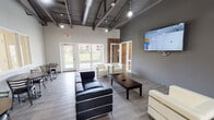 Located at 1958 W Algonquin Road in Mount Prospect, Illinois, this single-story amenity facility combines industrial-modern design with practical, community-focused spaces. The building's architecture is defined by soaring exposed steel roof trusses, visible ductwork, and pendant lighting throughout the main areas, creating an open, loft-inspired aesthetic that contrasts with clean white walls and light wood-look flooring.

The central lounge area serves as a welcoming gathering space, furnished with contemporary black leather and white upholstered seating arranged around a dark coffee table. A large wall-mounted display panel anchors the space, while glass-panel partitions and exterior glass doors flood the room with natural light and provide views of the surrounding green lawn.

Adjacent to the lounge, the fitness center is a generously sized, open workout space featuring rubber flooring, abundant natural light from a full bank of exterior windows, and a selection of cardio equipment including treadmills, stationary bikes, and elliptical machines. A dedicated flex room with exposed brick accent walls and hardwood-style flooring offers space for yoga or stretching. The facility also includes a well-appointed laundry room equipped with multiple stacked and top-load washers and dryers, finished with durable terrazzo-style flooring and bright overhead lighting.
