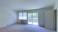 This single-floor space offers a clean and functional layout designed to maximize comfort and natural light. The main living area is highlighted by expansive windows and a full-width sliding glass door that floods the room with daylight and provides direct access to an outdoor deck, where views of lush, mature trees create a serene natural backdrop. The soft, wall-to-wall carpeting adds warmth and comfort underfoot, complementing the neutral, light-toned walls that keep the space feeling open and airy. A baseboard heating system runs along the perimeter of the room, ensuring efficient climate control throughout the colder months, while a wall-mounted air conditioning unit provides cooling during warmer seasons. The overall design is straightforward and versatile, offering a blank canvas that can easily be personalized to suit a variety of interior styles. The seamless connection between the interior living area and the outdoor deck enhances the sense of space, making it an ideal spot for relaxing or entertaining while enjoying the peaceful wooded surroundings.