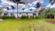 Located at 1920 Northeast 3rd Street in Deerfield Beach, Florida, this single-story property presents a classic Florida aesthetic characterized by a clean white exterior and a flat roofline. The building is set back from the street, surrounded by a generous expanse of well-maintained lawn that enhances its open, airy feel. Mature palm trees frame the property, lending it a distinctly tropical ambiance that is emblematic of South Florida living. Lush shrubs and landscaping line the base of the exterior walls, adding greenery and a manicured appearance to the grounds. A central air conditioning unit is visible along the exterior, ensuring year-round climate comfort. The building features multiple windows along its facade, allowing for natural light to enter the interior. The surrounding environment includes neighboring multi-story structures visible in the background, situating the property within an established and active neighborhood. The outdoor space is a defining feature of this property, offering a calm, verdant setting beneath the expansive Florida sky. Interior images were not sufficiently illuminated to provide detailed room-level descriptions.