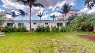 Located at 1920 Northeast 3rd Street in Deerfield Beach, Florida, this single-story property presents a classic Florida aesthetic characterized by a clean white exterior and a flat roofline. The building is set back from the street, surrounded by a generous expanse of well-maintained lawn that enhances its open, airy feel. Mature palm trees frame the property, lending it a distinctly tropical ambiance that is emblematic of South Florida living. Lush shrubs and landscaping line the base of the exterior walls, adding greenery and a manicured appearance to the grounds. A central air conditioning unit is visible along the exterior, ensuring year-round climate comfort. The building features multiple windows along its facade, allowing for natural light to enter the interior. The surrounding environment includes neighboring multi-story structures visible in the background, situating the property within an established and active neighborhood. The outdoor space is a defining feature of this property, offering a calm, verdant setting beneath the expansive Florida sky. Interior images were not sufficiently illuminated to provide detailed room-level descriptions.