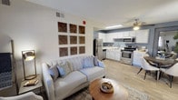 This single-floor residence offers a thoughtfully designed open-concept layout that seamlessly connects the living area to the kitchen. The living space features warm wood-tone flooring, a cozy fireplace with a classic metal screen, and a front entry door flanked by a window dressed in sheer white curtains that allow natural light to filter into the room. Decorative wall art composed of nine geometric wood panels adds a rustic, artisan touch to the neutral-toned walls, enhancing the welcoming ambiance of the space.

The kitchen is finished with crisp white cabinetry, light blue-gray accent walls, and a full suite of stainless steel appliances, including a refrigerator, microwave, and range. Light hardwood-style flooring continues throughout, creating a cohesive flow between both areas. A ceiling fan overhead provides comfort and airflow to the kitchen and dining vicinity.

The overall aesthetic is clean and contemporary, blending neutral tones with warm textures. The space is well-lit through a combination of natural light and overhead fixtures, making the interior feel open, bright, and livable.