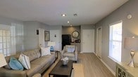 This single-floor space offers a clean, modern layout that combines a comfortable living area with a functional kitchenette in one open, cohesive environment. The living area is furnished with a plush sofa and matching armchair, centered around a dark coffee table and area rug, creating a welcoming and relaxed seating arrangement. Light hardwood flooring extends throughout the space, complementing the neutral gray walls and white trim for a contemporary, understated aesthetic. Recessed lighting in the ceiling provides bright, even illumination across the room. At the rear of the space, a compact kitchenette is equipped with a full-size black refrigerator, countertop appliances, and cabinetry with a warm wood finish, set within a defined nook that keeps the functional area neatly separated from the living zone. A large decorative wall clock serves as a focal point above the kitchenette counter, adding a stylish touch. Multiple doors along the walls suggest access to additional rooms or utility spaces. The textured ceiling and thoughtful layout contribute to a tidy, well-organized environment that balances comfort with practicality.