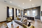 Located in Spring, TX, this single-floor combined kitchen and living room offers a contemporary and thoughtfully designed communal space. The living area features rich wood-look flooring, multiple seating arrangements with plush sofas and accent chairs in earthy tones, and a wall-mounted flat-screen display. Large windows dressed with dark drape panels invite generous natural light while offering views of the surrounding exterior. The kitchen area makes a strong design statement with its expansive white quartz island trimmed in deep navy cabinetry, complemented by a striking geometric patterned tile backsplash set against a dark accent wall. Bar-height seating lines the island, creating a natural gathering point. Sculptural pendant lighting in wire-cage and teardrop forms hang above, adding an artistic touch. Curated artwork adorns the walls throughout, reinforcing the space's modern aesthetic. Recessed lighting throughout ensures the entire space remains bright and welcoming. A secondary lounge nook with a wall-mounted screen and additional seating provides a more intimate setting. The seamless flow between the kitchen and living zones makes this a highly functional and visually cohesive space suited for gathering and entertaining.