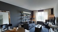 This single-floor space showcases a thoughtfully designed living area defined by its bold, dark slate-grey walls that create a dramatic yet refined atmosphere. The room is anchored by a charcoal button-tufted sofa adorned with accent pillows, complemented by a dark upholstered armchair nearby. A striking open metal-and-white shelving unit serves as a focal point along one wall, displaying decorative objects, small plants, and curated accessories that add personality to the space. A low, dark coffee table sits atop a deep-toned area rug, grounding the seating arrangement. Natural light filters generously through a large window dressed with sheer white curtains and a dark Roman shade, offering views of the surrounding greenery and street below. The contrast between the bright white ceiling and the deep wall color adds vertical dimension to the room. Warm-toned table lamps provide ambient lighting, softening the overall mood. A round dining or accent table with a woven tray detail is partially visible in the foreground, suggesting additional functional space. The overall ambiance is sophisticated, urban, and cohesive, with a carefully curated mix of textures and tones throughout.