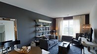 This single-floor space showcases a thoughtfully designed living area defined by its bold, dark slate-grey walls that create a dramatic yet refined atmosphere. The room is anchored by a charcoal button-tufted sofa adorned with accent pillows, complemented by a dark upholstered armchair nearby. A striking open metal-and-white shelving unit serves as a focal point along one wall, displaying decorative objects, small plants, and curated accessories that add personality to the space. A low, dark coffee table sits atop a deep-toned area rug, grounding the seating arrangement. Natural light filters generously through a large window dressed with sheer white curtains and a dark Roman shade, offering views of the surrounding greenery and street below. The contrast between the bright white ceiling and the deep wall color adds vertical dimension to the room. Warm-toned table lamps provide ambient lighting, softening the overall mood. A round dining or accent table with a woven tray detail is partially visible in the foreground, suggesting additional functional space. The overall ambiance is sophisticated, urban, and cohesive, with a carefully curated mix of textures and tones throughout.