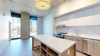 Located in the heart of downtown Kansas City, this contemporary single-floor residence at 81 E 14th St delivers an striking blend of industrial character and modern sophistication. Exposed concrete ceilings and structural columns provide an industrial-chic aesthetic that contrasts beautifully with the clean white walls and light hardwood flooring found throughout the space.

The open-concept kitchen is a standout feature, outfitted with stainless steel appliances including a French door refrigerator, built-in microwave, dishwasher, and range. Sleek two-toned cabinetry pairs white upper cabinets with warm wood lower cabinets, complemented by a marble-look tile backsplash. A large center island with a white quartz countertop provides ample workspace and additional storage below.

The living area is defined by dramatic floor-to-ceiling windows that frame sweeping panoramic views of the Kansas City skyline, flooding the space with abundant natural light. Exposed concrete columns add architectural interest throughout.

The bedroom offers a generous layout with direct access to a walk-in closet and an en-suite bathroom. The bathroom is finished with marble-look tile on both the floors and walls, a glass-enclosed walk-in shower, a dual-sink vanity with white countertops, and modern chrome fixtures — creating a clean, spa-like atmosphere.