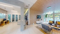 This thoughtfully designed single-floor amenity space combines contemporary aesthetics with practical functionality across several distinct zones. The central lounge area serves as the heart of the space, featuring a striking wood-paneled accent wall and ceiling with a large mounted television, modern gray and yellow upholstered seating, and an area rug anchoring the arrangement. Floor-to-ceiling glazing offers sweeping views of a resort-style outdoor pool framed by lush palm trees, flooding the interior with natural light.

Adjacent to the lounge, a sleek communal kitchen features white cabinetry, wood accents, a waterfall-edge island with seating, pendant lighting, and a full wall of glass overlooking the tropical outdoor setting. The business center offers a long yellow workstation with multiple computer stations, pendant lighting, and generous window views of greenery. Glass-enclosed private meeting rooms provide semi-transparent privacy and are visible from the main work area, alongside a multifunction printer station.

The fitness center is well-appointed with cardio equipment, cable machines, free weights, and a mirrored wall, all illuminated by abundant natural light through large windows overlooking the pool deck. Large-format white tile flooring, recessed lighting, and a clean white palette unify the overall space.