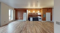 This single-floor space offers a versatile, open-plan layout finished with light wood-look flooring that runs throughout, lending warmth and visual continuity to the entire unit. The main living area is generously proportioned, with clean white walls and exposed ceiling-mounted conduit and track elements that give the space a subtle industrial character. A wall-mounted HVAC unit ensures climate control comfort.

The kitchen area makes a bold design statement with rich terracotta and burnt orange accent walls that contrast sharply against the dark lower cabinetry and neutral gray countertops. Open wall-mounted shelving and upper cabinets provide practical storage, while three globe pendant lights hang overhead, creating a warm, focused ambiance. The kitchen is fully equipped with a white electric range, dishwasher, refrigerator, and a single-basin sink, making it functional for everyday use.

A closet with double panel doors and an electrical panel are neatly integrated into the kitchen wall. Multiple entry points, including two separate doors along the main wall, offer flexible access to the space. The floor plan, as revealed in the overhead dollhouse view, flows in an open L-shaped configuration, maximizing usable floor area and providing ample room for varied furnishing arrangements. Overall, the space balances industrial details with warm, inviting finishes.