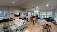 Located at 2200 Brown St in Waxahachie, TX, this single-story community clubhouse at BridgePoint offers a well-appointed, open-concept gathering space designed for both relaxation and productivity. The interior is anchored by warm hardwood-style flooring that runs throughout, complemented by vaulted ceilings that lend an airy, expansive feel to the space.

The main lounge area features comfortable seating arrangements, including a gray sofa adorned with colorful accent pillows, grouped around patterned area rugs. A teal-accented wall frames a tile-surround fireplace, creating a welcoming focal point for residents to gather. Framed artwork is thoughtfully displayed throughout, adding a polished, curated aesthetic.

Adjacent to the lounge, a stylish bar-height island with a rich butcher-block wood top serves as a social hub, illuminated by modern geometric pendant lighting. Large windows fitted with blinds line the walls, flooding the space with natural light and offering views of the surrounding greenery.

A dedicated business center includes a long wooden counter with computer workstations and wire bar stools, positioned beneath vaulted ceilings and a generous wall of windows. Bistro-style tables and wire-frame chairs are scattered throughout for flexible seating. Private office spaces visible toward the rear of the clubhouse provide additional functionality for leasing and administrative use.