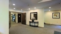 This single-floor space presents a well-appointed corridor or common hallway area designed with a clean, understated aesthetic. The space is illuminated by a series of recessed ceiling lights that cast an even, welcoming glow throughout the area, complementing the neutral-toned walls and textured wall-to-wall carpeting underfoot. The carpet features a subtle pattern in muted tones, contributing to the overall sense of quiet sophistication.

A standout decorative feature along one wall is a dark-framed rectangular mirror hung above a curved, dark metal console table. The table is accented with a dark ceramic vase holding dried floral stems, adding a refined, tasteful touch to the space. This vignette creates a focal point that lends warmth and character to what might otherwise be a purely functional corridor.

A solid dark wood door with hardware is visible to one side, suggesting secure access to an adjacent area, while a glimpse through an open passage hints at additional space beyond. The ceiling features a smooth finish with a textured border detail, adding a subtle architectural element. Overall, the space conveys a tidy, well-maintained environment with a calm and welcoming ambiance suited to a shared or transitional interior setting.