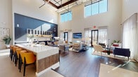Located at 305 Prine Pl in Charlotte, NC, this single-story community clubhouse — branded under the "Well | Live + Thrive" concept — is a thoughtfully designed gathering space that blends modern architecture with warm, welcoming interiors. The entrance greets visitors with polished concrete floors, black-framed glass double doors, and a curated gallery wall, setting an refined tone from the moment you arrive. The main lounge area opens into a dramatic, double-height space bathed in natural light from expansive floor-to-ceiling windows dressed with sheer linen drapery. Rich hardwood flooring extends throughout, complementing a sophisticated palette of navy, warm wood tones, and crisp white. A striking central staircase with steel cable railings rises against a wall-to-wall, floor-to-ceiling built-in bookcase with integrated lighting, serving as a true architectural focal point. A long bar counter with bold yellow upholstered barstools anchors the social hub of the space. The kitchenette features sleek slate-blue cabinetry, quartz countertops, open floating shelves, and stainless steel appliances. Multiple seating arrangements — including plush sofas, accent chairs, and coffee tables — encourage both casual conversation and relaxation. A wall-mounted television and ample lounge seating complete this polished, community-forward environment.