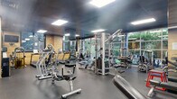 Located at 100 Paterson Plank Rd in Jersey City, NJ, this fitness center offers a comprehensive, single-floor layout designed to support a full range of wellness activities. The main exercise room is generously proportioned, featuring durable rubber flooring, a dark paneled ceiling with recessed panel lighting, and warm wood-accent walls that lend the space a modern yet inviting atmosphere. Floor-to-ceiling glass windows along one side of the gym flood the area with natural light and offer views of the surrounding outdoor greenery. The facility is outfitted with an extensive array of equipment, including treadmills, elliptical machines, cable and strength machines, free weights, and adjustable benches. Large mirrors line the walls to enhance the sense of space and support training. A wall-mounted television provides entertainment during workouts. Adjacent to the main gym area is a dedicated relaxation lounge, enclosed by frosted glass partitions and featuring lounge chairs with ottomans set against a light wood accent wall — an ideal space for post-workout recovery. The facility also includes a traditional wood-paneled sauna, complete with tiered benches, a Himalayan salt wall feature, and a small window for natural light, creating a warm and restorative retreat.