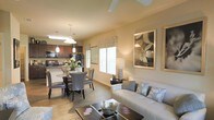 This single-floor residence showcases an open-concept floor plan that effortlessly integrates the living, dining, and kitchen areas into one cohesive, welcoming space. The living room is anchored by a comfortable neutral-toned sofa accented with decorative pillows, complemented by a stylish coffee table and tasteful wall art that adds a sophisticated touch. Natural light pours in through multiple windows dressed with decorative valances, creating a warm and inviting ambiance throughout. A ceiling fan overhead adds both function and a casual elegance to the space. The dining area features a round glass-top table surrounded by upholstered chairs, illuminated by a pendant light fixture that adds a refined focal point. The kitchen, visible from the dining area, is equipped with dark cabinetry, black appliances, and a breakfast bar that enhances both its functionality and open feel. Pendant lighting above the kitchen counter echoes the design choices found throughout the home. Warm wood-look flooring runs continuously through the main living areas, tying the spaces together and adding a sense of warmth and continuity. The overall aesthetic is modern yet approachable, with a neutral color palette and carefully chosen furnishings that make this home feel both stylish and comfortable.