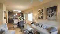 This single-floor residence showcases an open-concept floor plan that effortlessly integrates the living, dining, and kitchen areas into one cohesive, welcoming space. The living room is anchored by a comfortable neutral-toned sofa accented with decorative pillows, complemented by a stylish coffee table and tasteful wall art that adds a sophisticated touch. Natural light pours in through multiple windows dressed with decorative valances, creating a warm and inviting ambiance throughout. A ceiling fan overhead adds both function and a casual elegance to the space. The dining area features a round glass-top table surrounded by upholstered chairs, illuminated by a pendant light fixture that adds a refined focal point. The kitchen, visible from the dining area, is equipped with dark cabinetry, black appliances, and a breakfast bar that enhances both its functionality and open feel. Pendant lighting above the kitchen counter echoes the design choices found throughout the home. Warm wood-look flooring runs continuously through the main living areas, tying the spaces together and adding a sense of warmth and continuity. The overall aesthetic is modern yet approachable, with a neutral color palette and carefully chosen furnishings that make this home feel both stylish and comfortable.